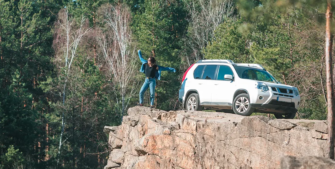 Lady standing on a cliff with an SUV behind her.