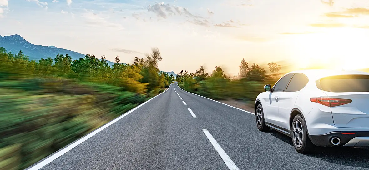 A car driving down a tree lined, winding road into the sunset.