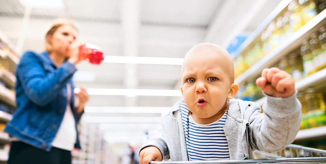 Baby in supermarket basket seat with fist raised.