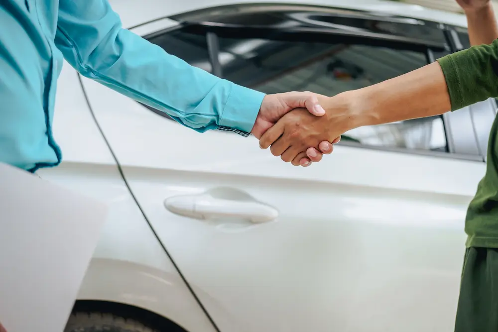 Two people stand in front of a car shaking hands, only their arms and hands are visible in the frame. They stand in front of the back door of a white car.