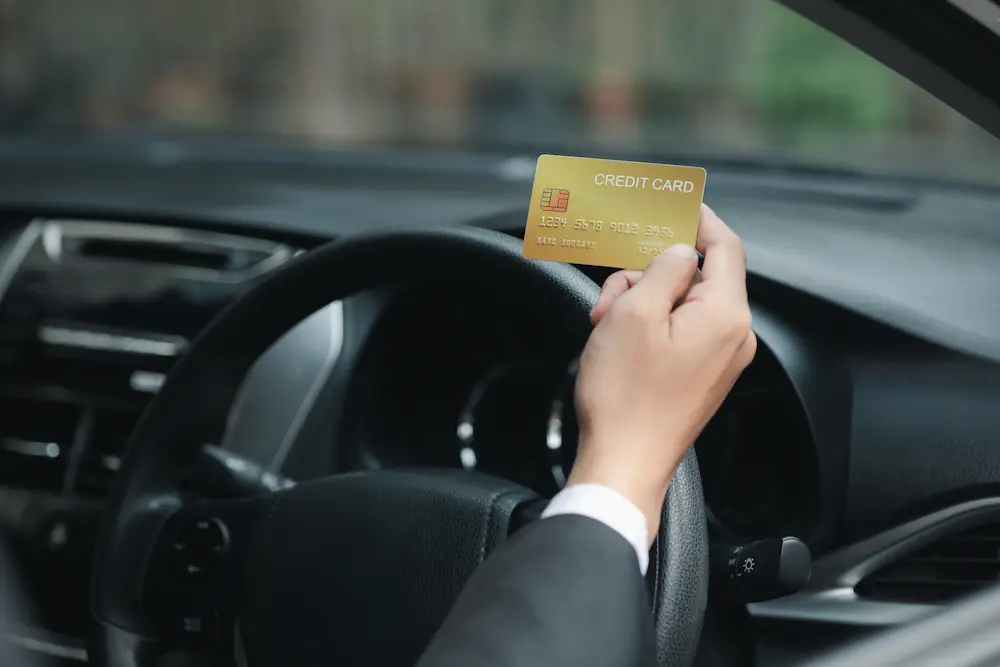 A man's hand hovers above a black steering wheel holding a gold credit card. The interior of the car is black and the man is wearing a black suit jacket.