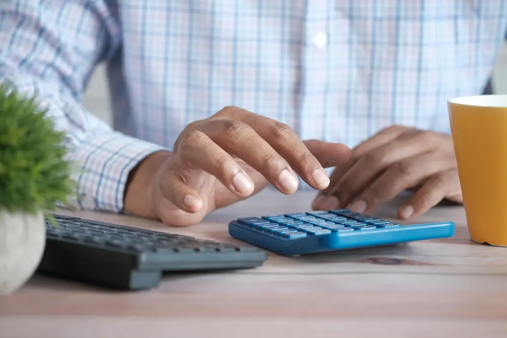 A person using a blue calculator on a wooden desk with a keyboard and plant nearby