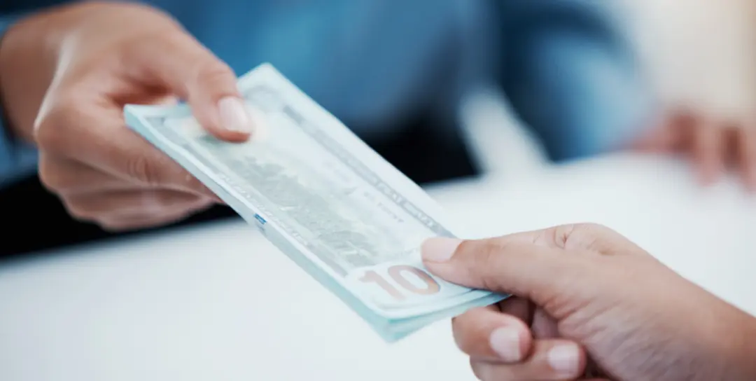 Close-up on the hands of one person handing a stack of $100 bills to another person across a white table.
