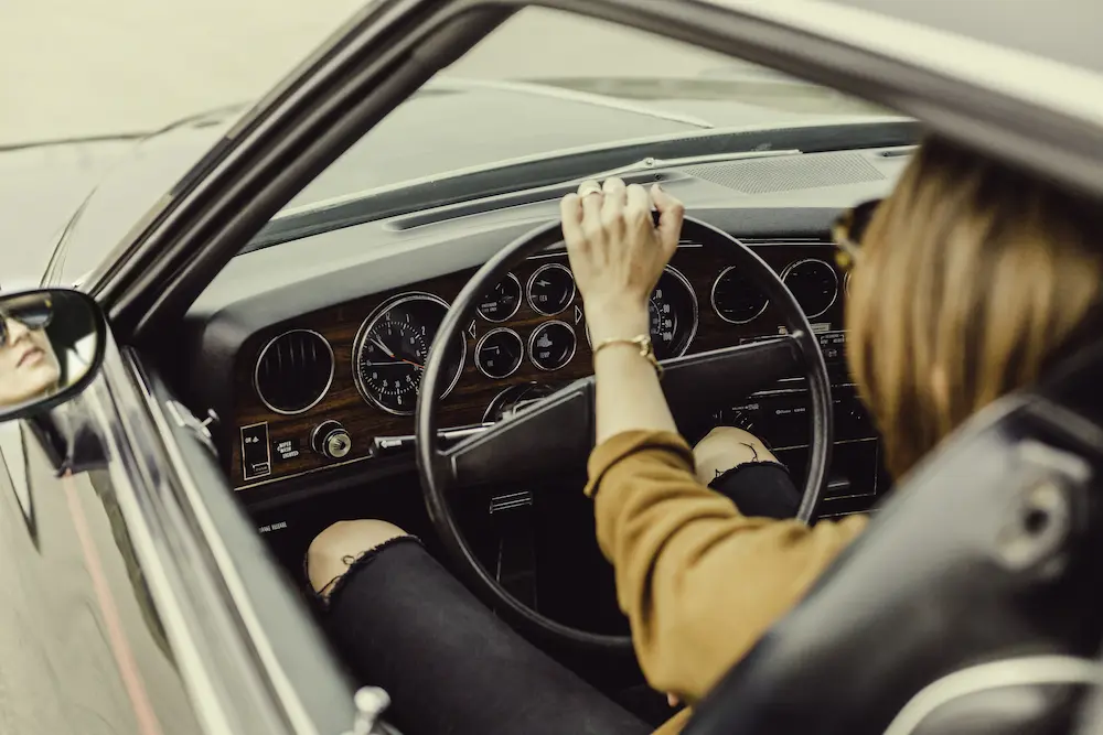 View from behind the driver side door of a young driver behind the wheel of a dark car wearing a brown sweater and ripped black jeans