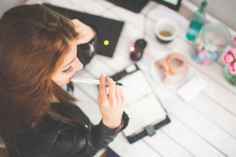 Overhead view of a brunette woman working at a desk resting a pen on her chin with a planner open and items on her desk in the background