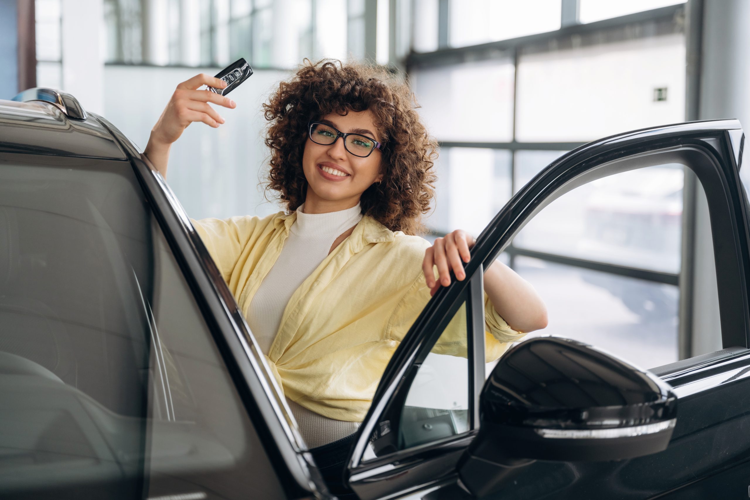 Young woman with glasses and curly hair stands in the open door of a car, holding up a keyfob.