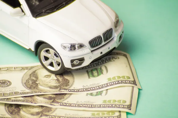 A toy car sits atop several one-hundred dollar bills, set against a green background.